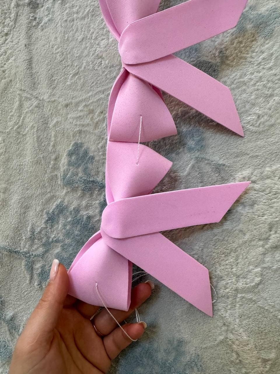 May include: A light pink ribbon made of foam, with several loops and pointed ends. The ribbon is sewn together with white thread. The ribbon is held by a hand with a light-colored manicure.