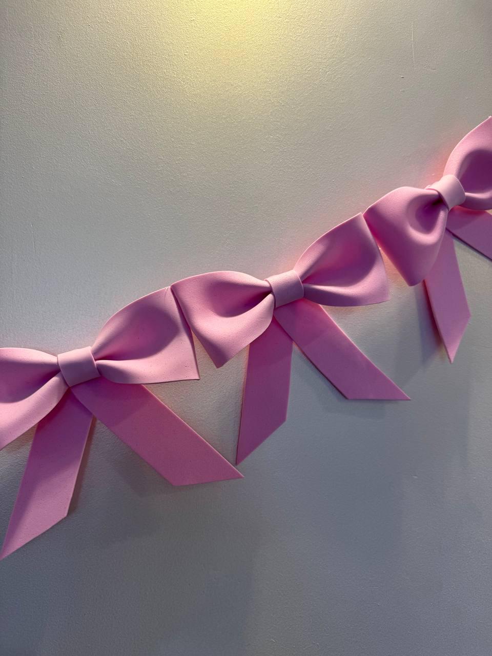 May include: A series of pink bows are arranged diagonally against a light gray wall. The bows are a solid, soft pink color and appear to be made of a smooth material. The bows are evenly spaced and create a decorative pattern.