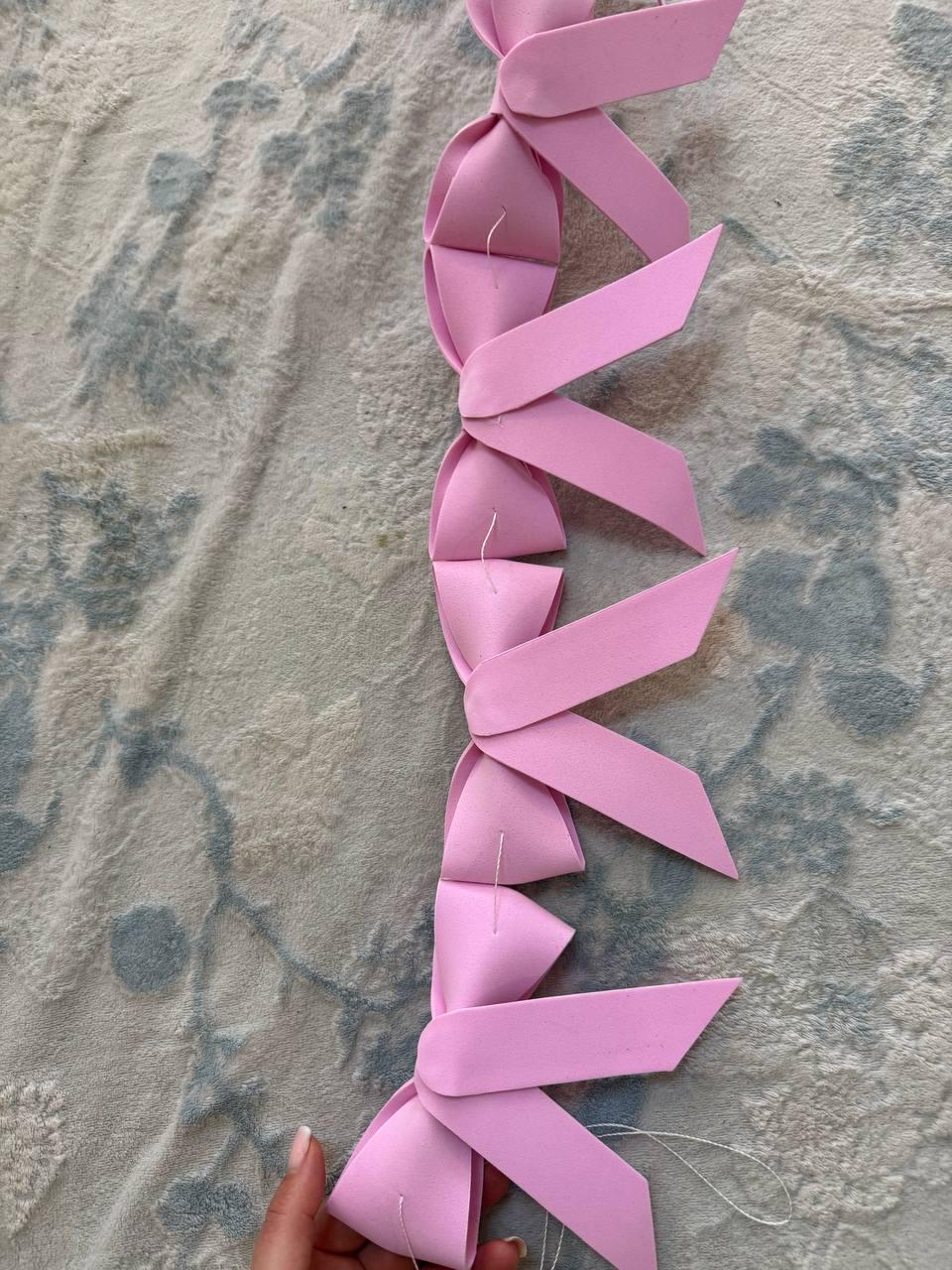 May include: A garland of pink paper bows, arranged in a vertical chain. The bows are made of folded paper and are connected with white thread. The garland is displayed against a patterned, light-colored fabric background.
