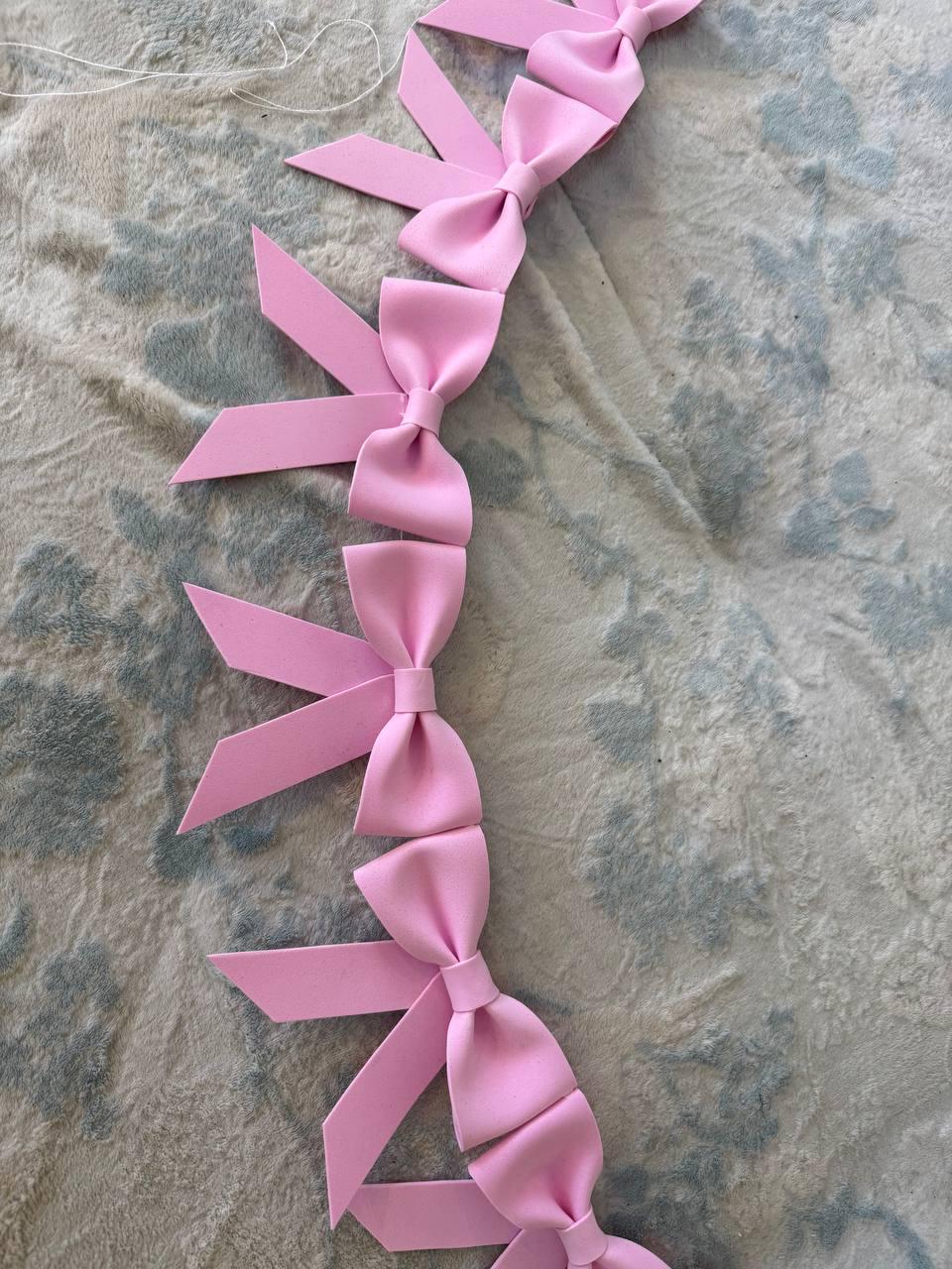 May include: A string of light pink bows, each with a looped center and long, pointed tails. The bows are evenly spaced and connected by a thin white string, creating a garland or decorative element. The bows are made of a soft material.