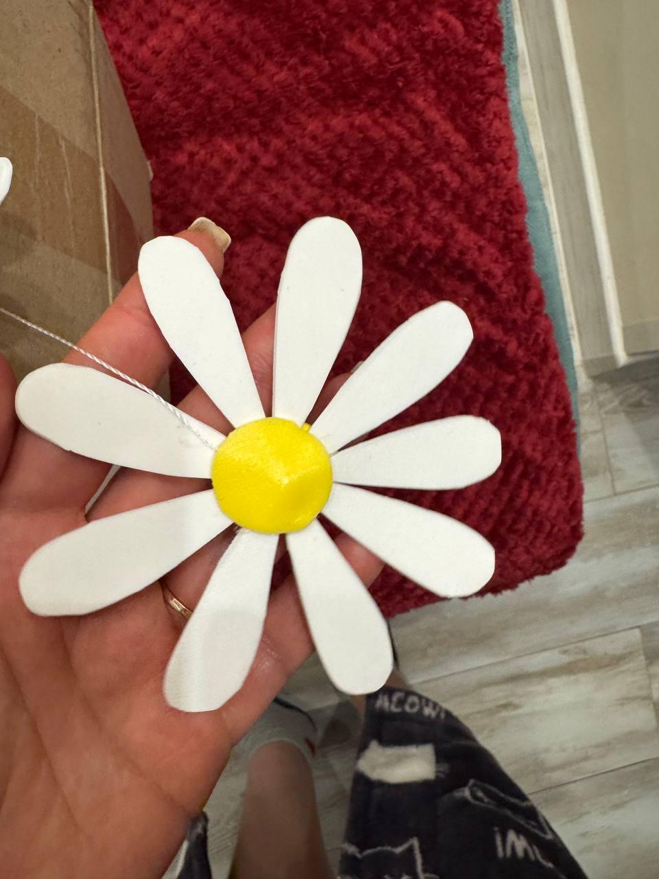 May include: A large, handmade daisy with white petals and a yellow center. The flower is held against a red textured background. The daisy is made of a lightweight material and has a string attached for hanging or crafting.