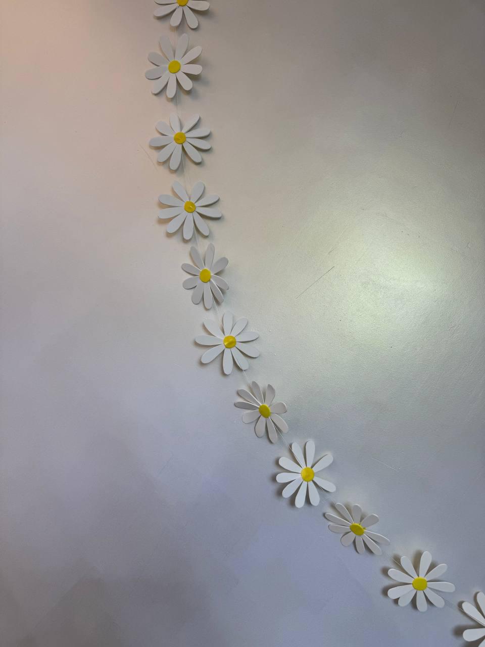 May include: A garland of white paper daisies with yellow centers. The daisy chain is strung diagonally against a white background. The daisy petals are layered, giving a 3D effect.