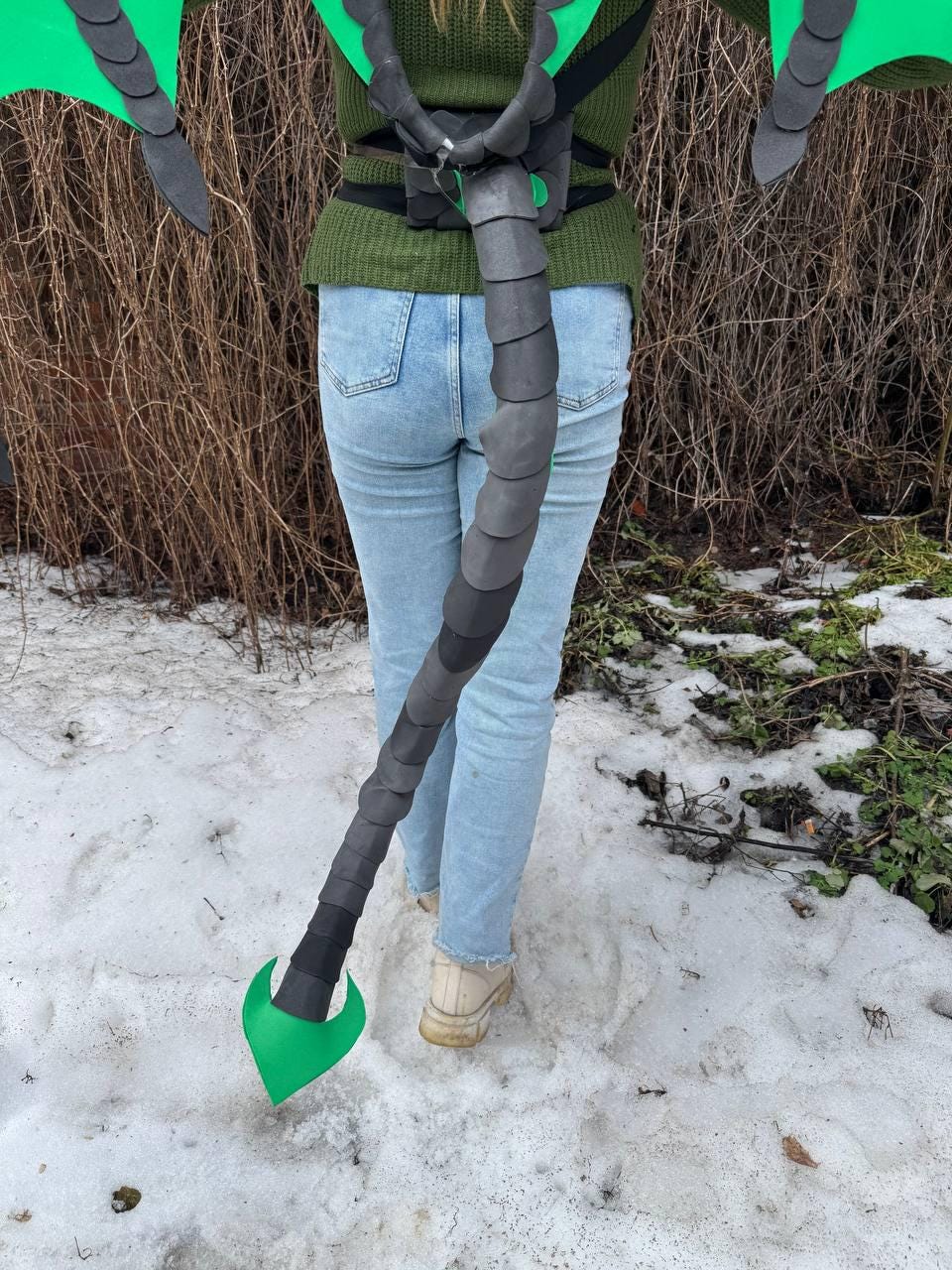 May include: A black and gray dragon tail prop with green accents. The tail is attached to a black harness and is worn by a person wearing a green sweater and blue jeans. The tail is made of layered, curved segments.