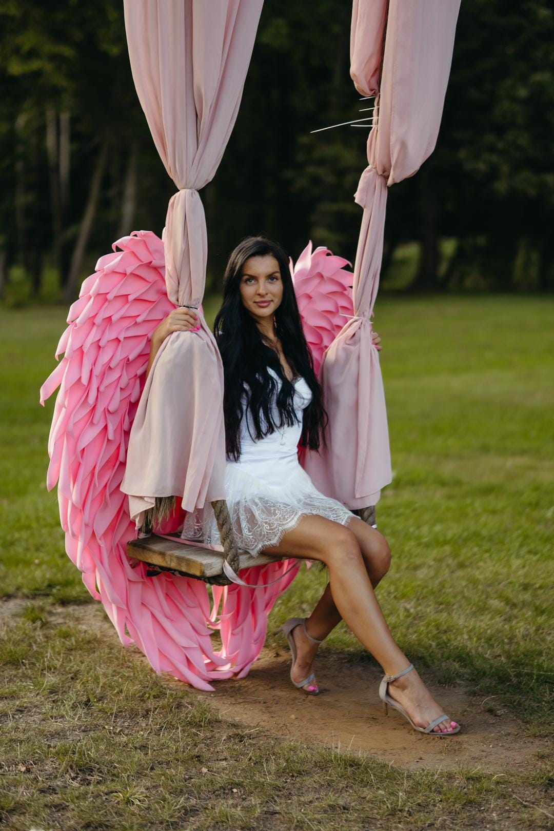 May include: A woman sits on a wooden swing adorned with large, pink feathery wings. The swing is suspended by pink fabric from above. She wears a white dress with lace trim and strappy heels, set against a backdrop of green grass and trees.
