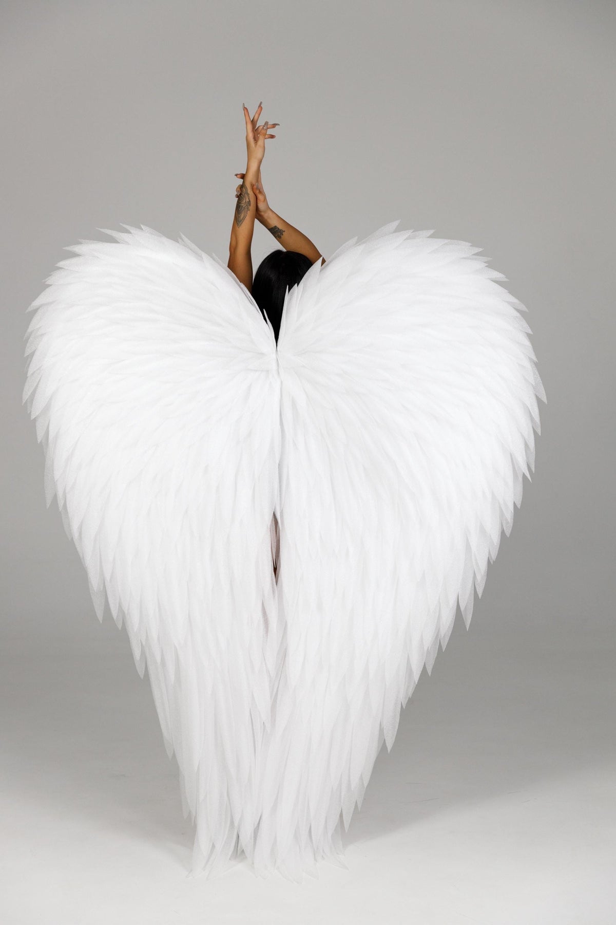 May include: Large, white feathered angel wings. The wings are spread wide, with a heart-like shape. The feathers are layered and detailed. The person's arms are raised above their head.