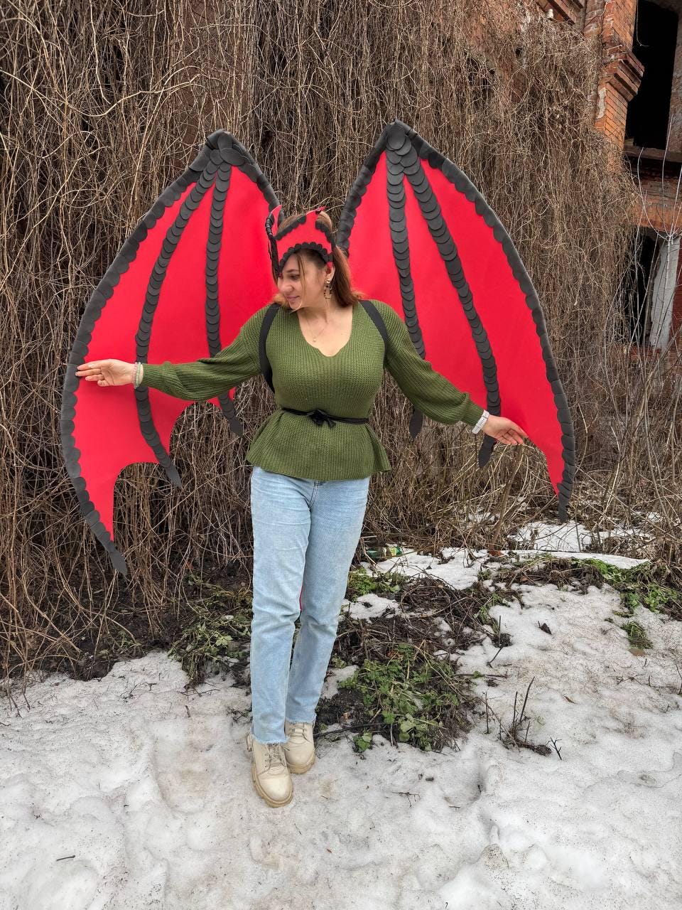 May include: A person wearing a green sweater and blue jeans, with large red and black dragon wings and a matching headband. The wings are spread wide, showcasing their vibrant color against a backdrop of winter foliage and snow.