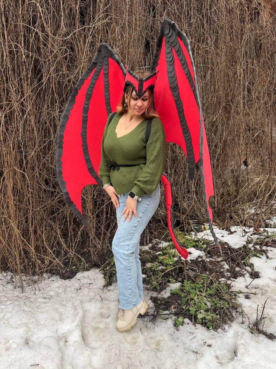 May include: A red and black dragon wing costume with a matching headpiece. The wings are large and detailed, with a red fabric and black trim. The headpiece is red and black, with a pointed design. The costume is worn with a green sweater and blue jeans.
