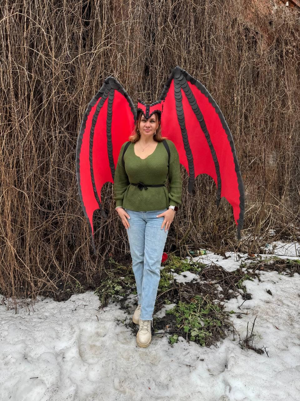 May include: A person wearing a green sweater, blue jeans, and beige boots, posing in front of a backdrop of bare branches. The person is wearing a red and black dragon wing costume, with a matching headpiece.
