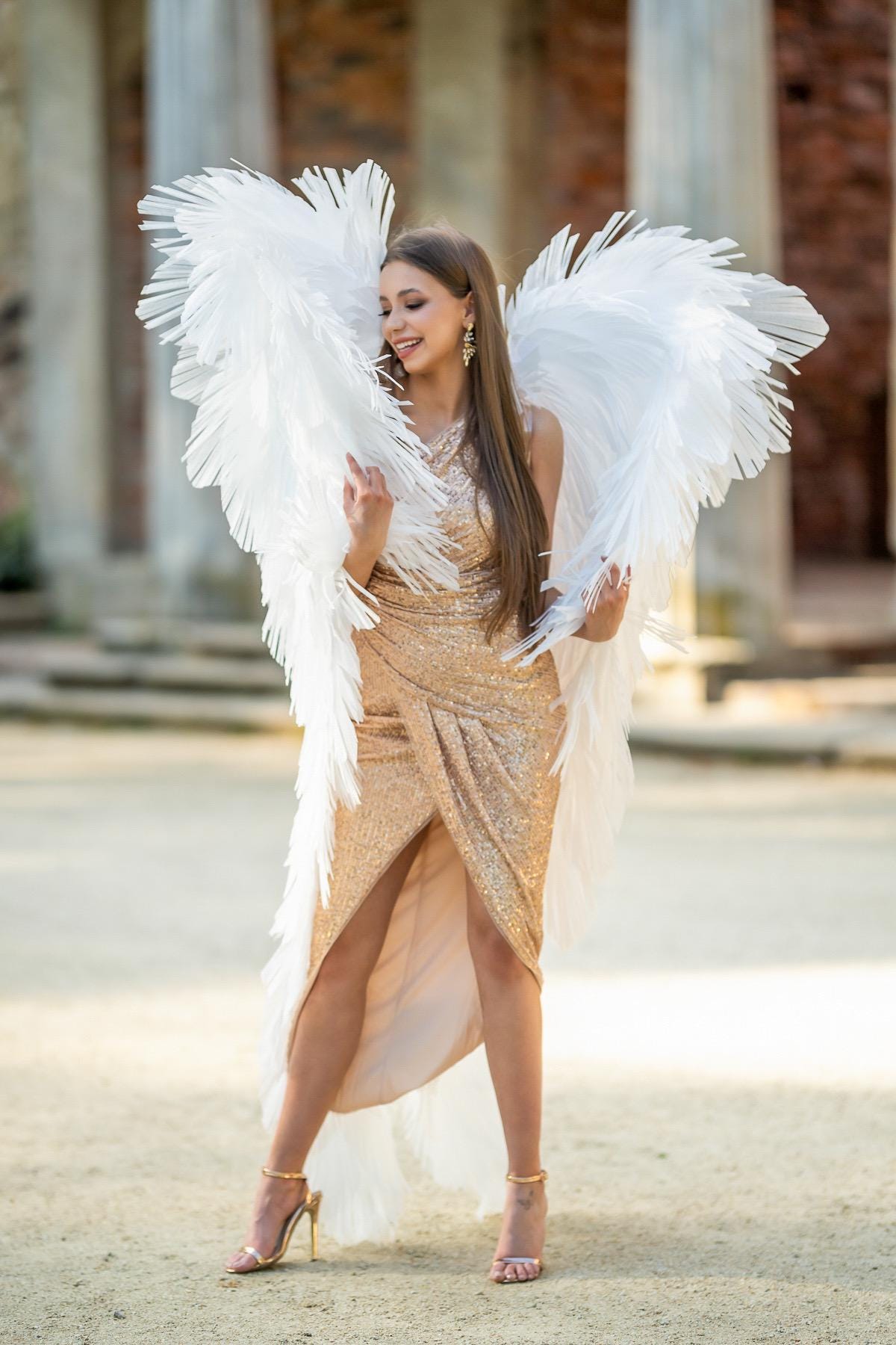May include: A woman wearing a gold sequined dress and large white feather angel wings. The dress has a wrap design with a high slit. She is wearing gold heels. The background features stone columns and a paved area.