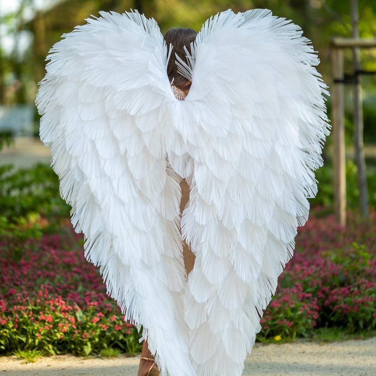 May include: Large, white feather angel wings. The wings are made of many individual feathers, creating a full, textured appearance. The wings are worn by a person, and the image is taken from behind, showcasing the wings' impressive size and design.