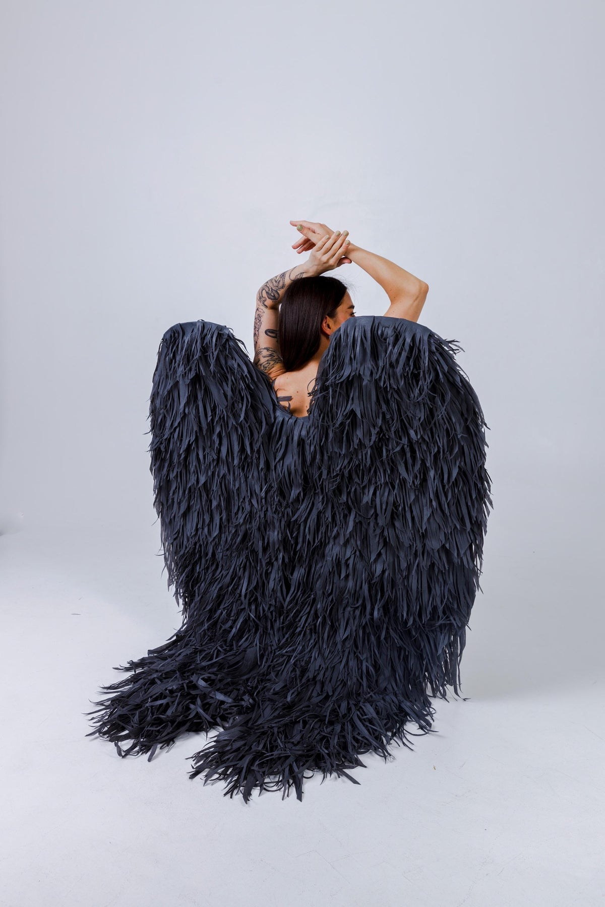 May include: A pair of large, dark gray angel wings with a feathered texture. The wings are worn on the back, with the wearer's arms raised. The wings are made of a soft, flowing material.
