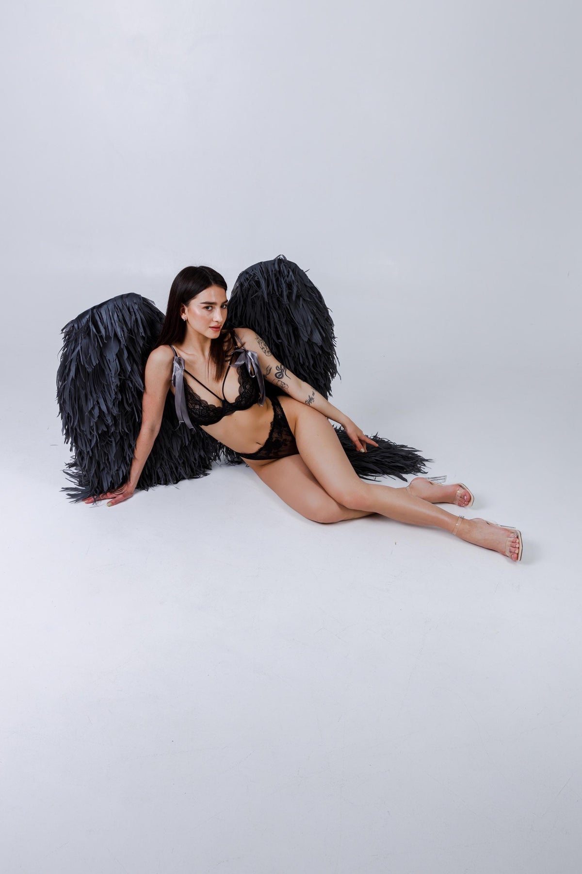 May include: A black lingerie set with lace detailing and a matching pair of large, feathered angel wings. The model is wearing the lingerie and wings, and is lying on a white surface.