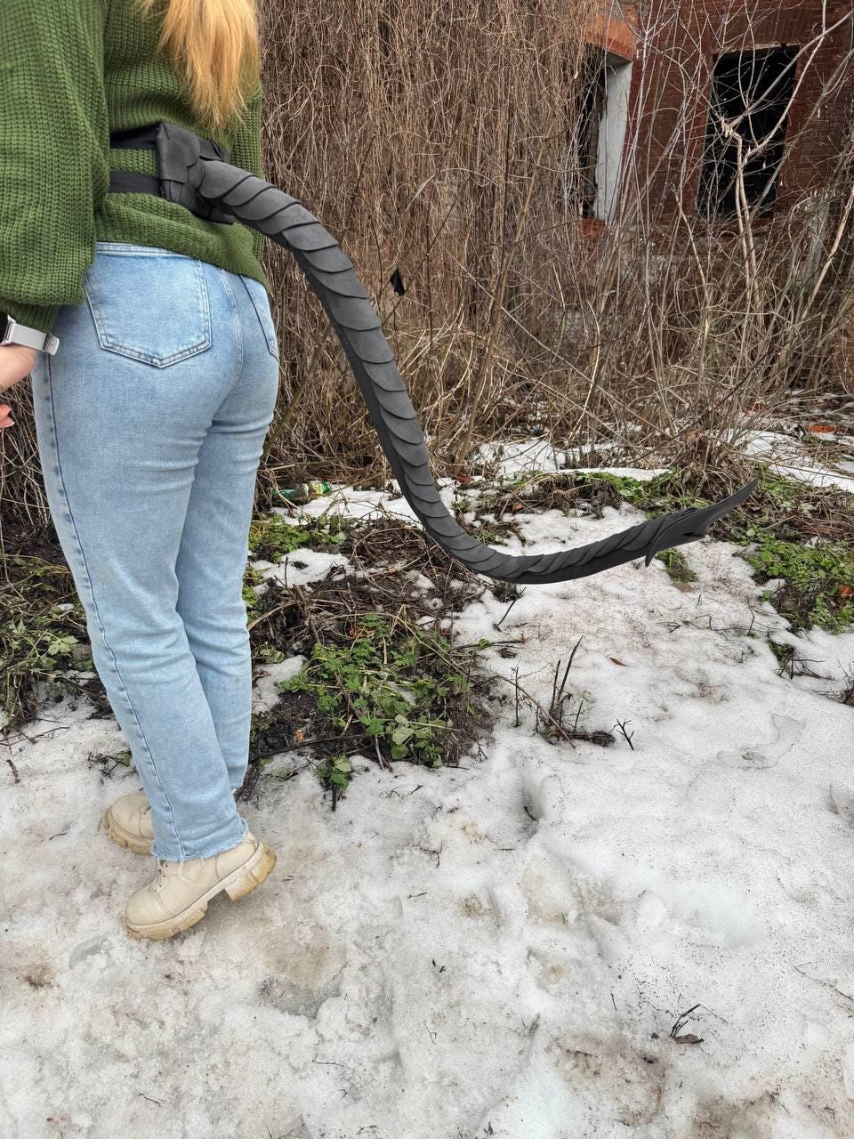 May include: A black, segmented tail prop, attached to a black belt, curves downward. The tail is worn by a person wearing a green sweater and light blue jeans. The background features snow, grass, and bare branches.
