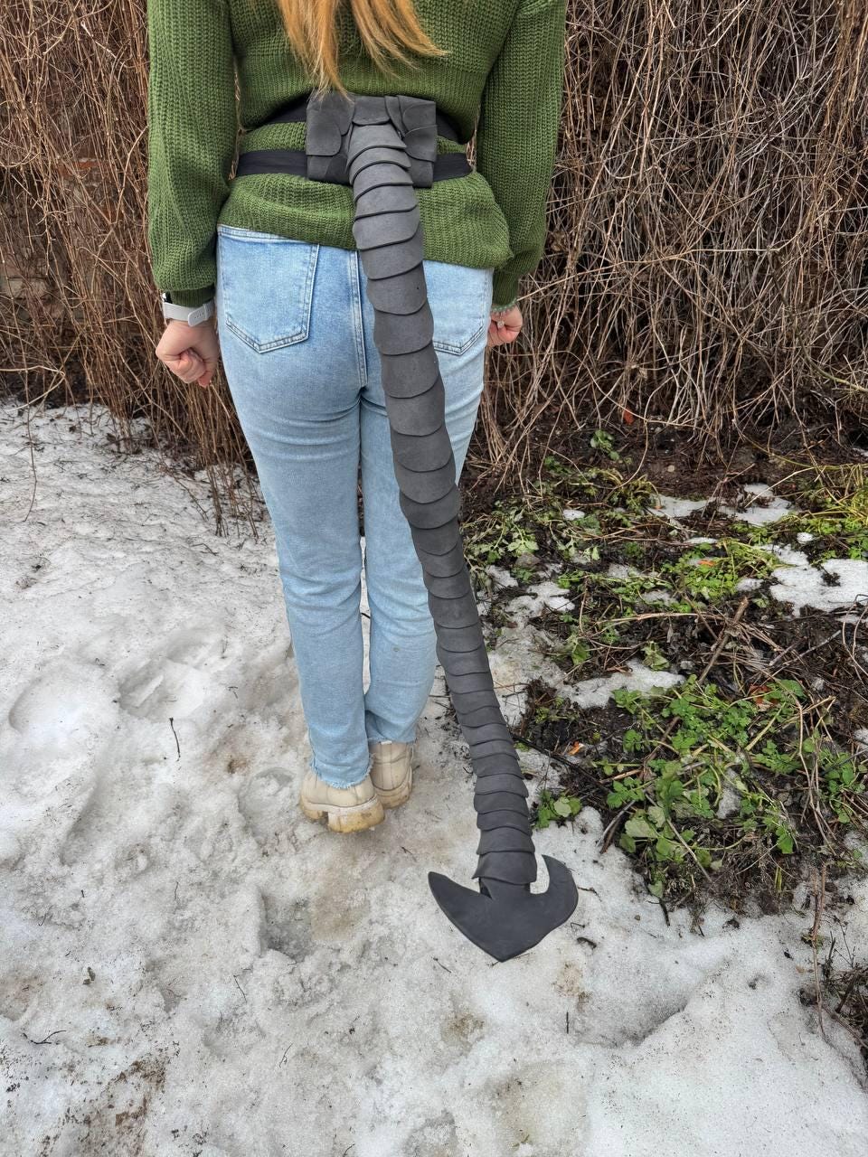 May include: A black, segmented tail prop, attached to a black belt, hangs down. The tail is made of a dark gray material and has a curved, pointed end. The belt is worn with light blue jeans and a green sweater.
