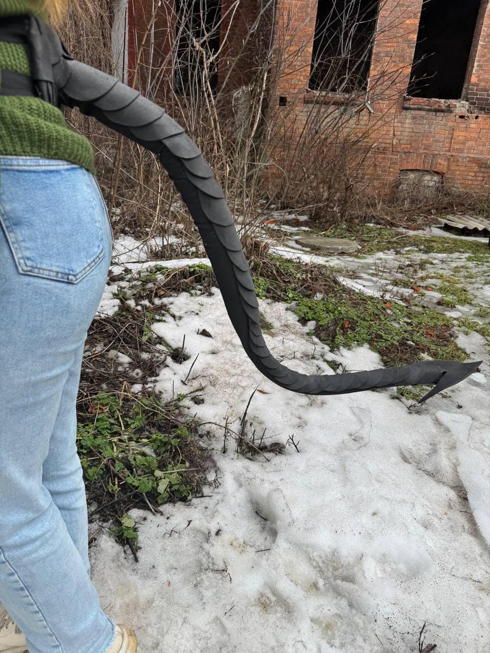 May include: A black, scaled tail prop, likely for cosplay or costume use. The tail is curved and flexible, with a pointed tip. The tail is attached to a harness, and the person is wearing a green sweater and blue jeans.