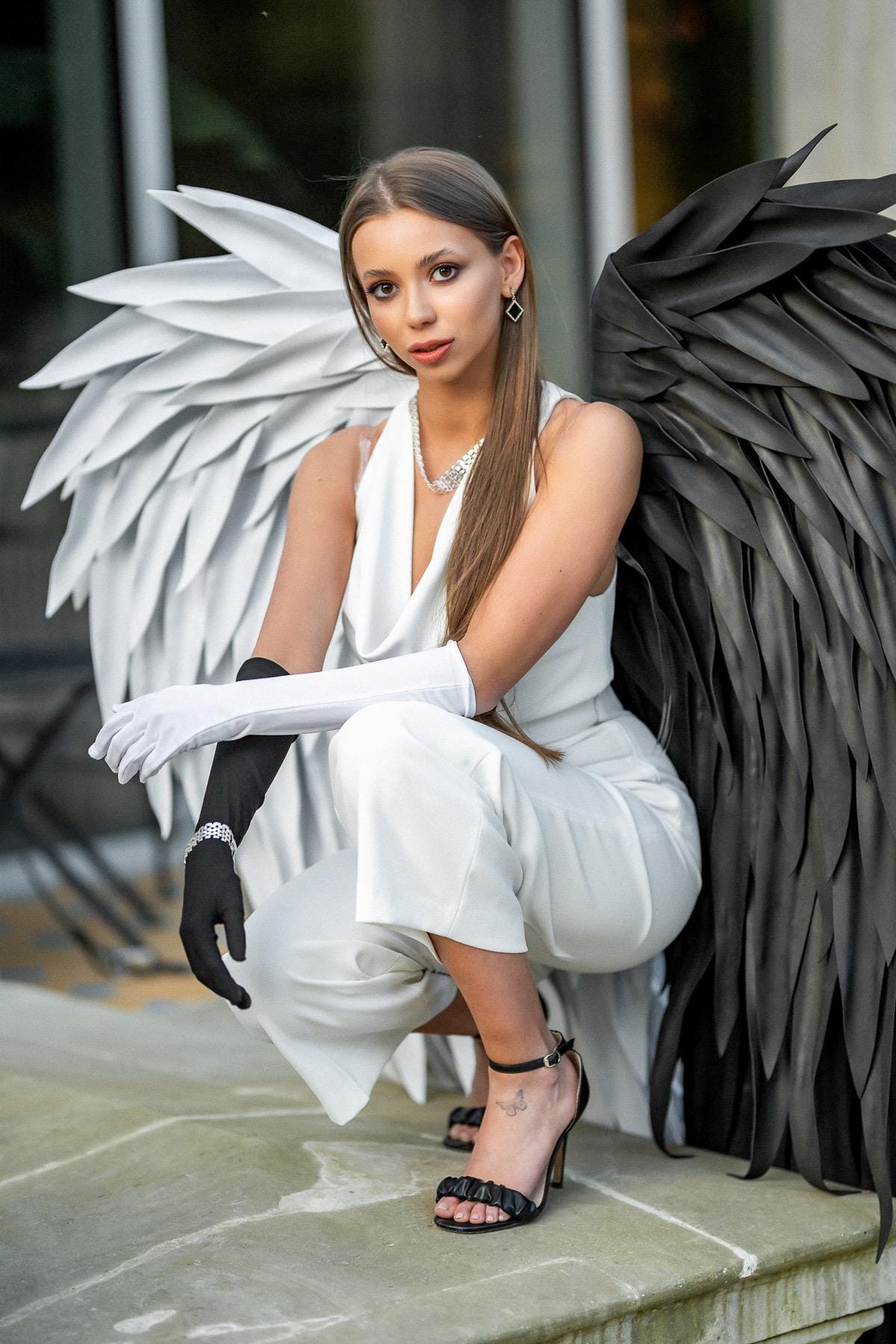 May include: A white sleeveless jumpsuit with a cowl neckline, paired with one white opera glove and one black opera glove. Large, layered white wings extend from the right side and large, layered black wings extend from the left side.