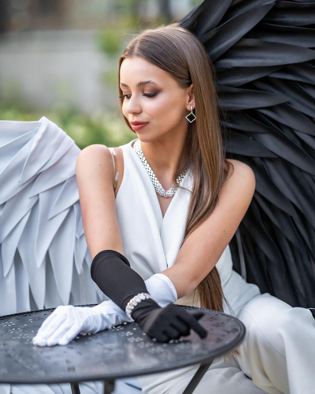 May include: A white sleeveless jumpsuit paired with long black gloves, a silver chain necklace, and square drop earrings. Large white and black feathered wings are positioned behind the shoulders, creating a contrast between the light and dark elements.