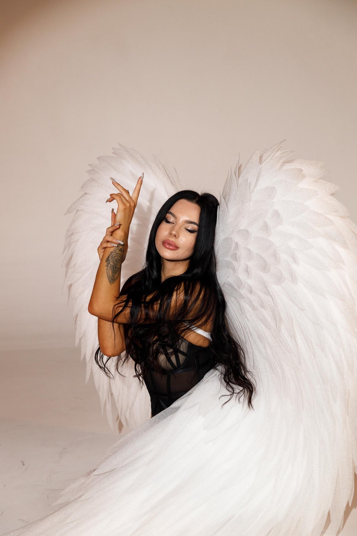 May include: A woman with long black hair and large white feathered wings. She is wearing a black top. The background is a light beige color.