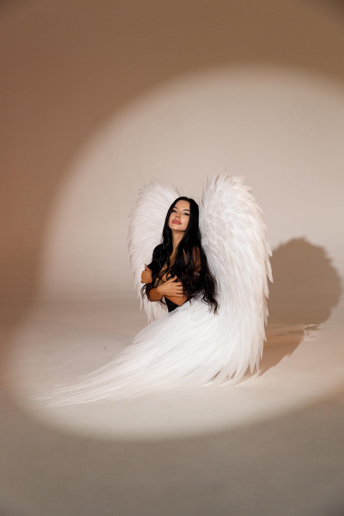 May include: A person wearing a black top and large white feathered angel wings. The wings are spread out behind the person, creating a dramatic effect. The background is a neutral color.