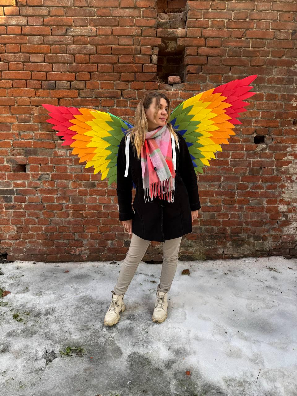 May include: A person wearing large, colorful rainbow-colored wings stands in front of a brick wall. The wings feature red, orange, yellow, green, and blue hues. The person is wearing a black coat, tan pants, and white boots.