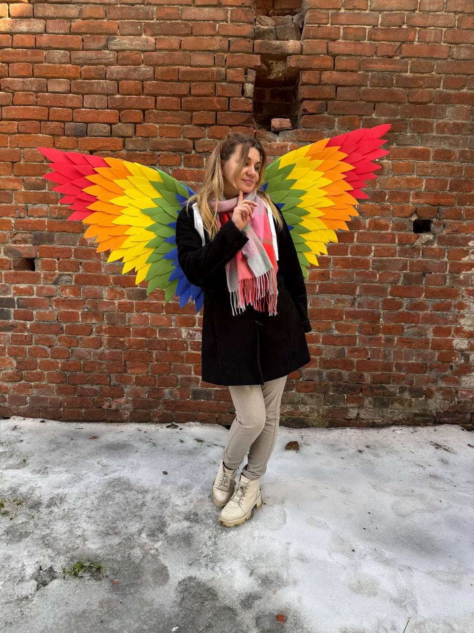 May include: A person wearing colorful rainbow-colored wings, standing in front of a brick wall. The wings are made of layered, colorful material, with red, orange, yellow, green, and blue hues. The person is wearing a black coat, beige pants, and white boots.