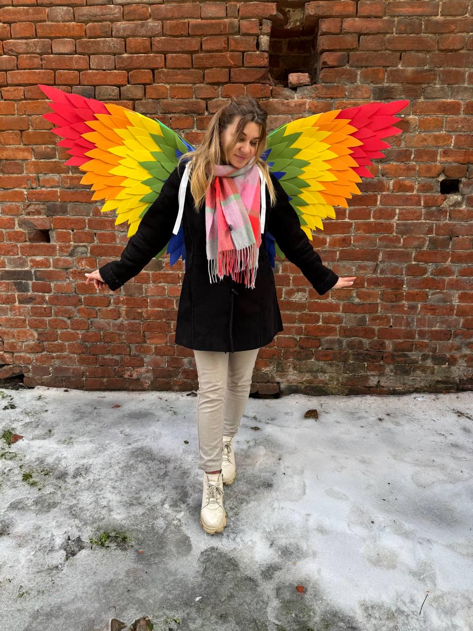 May include: A person wearing colorful rainbow-colored wings, a black coat, beige pants, and white boots. The wings are made of layered, colorful felt in red, orange, yellow, green, and blue. The background is a brick wall.