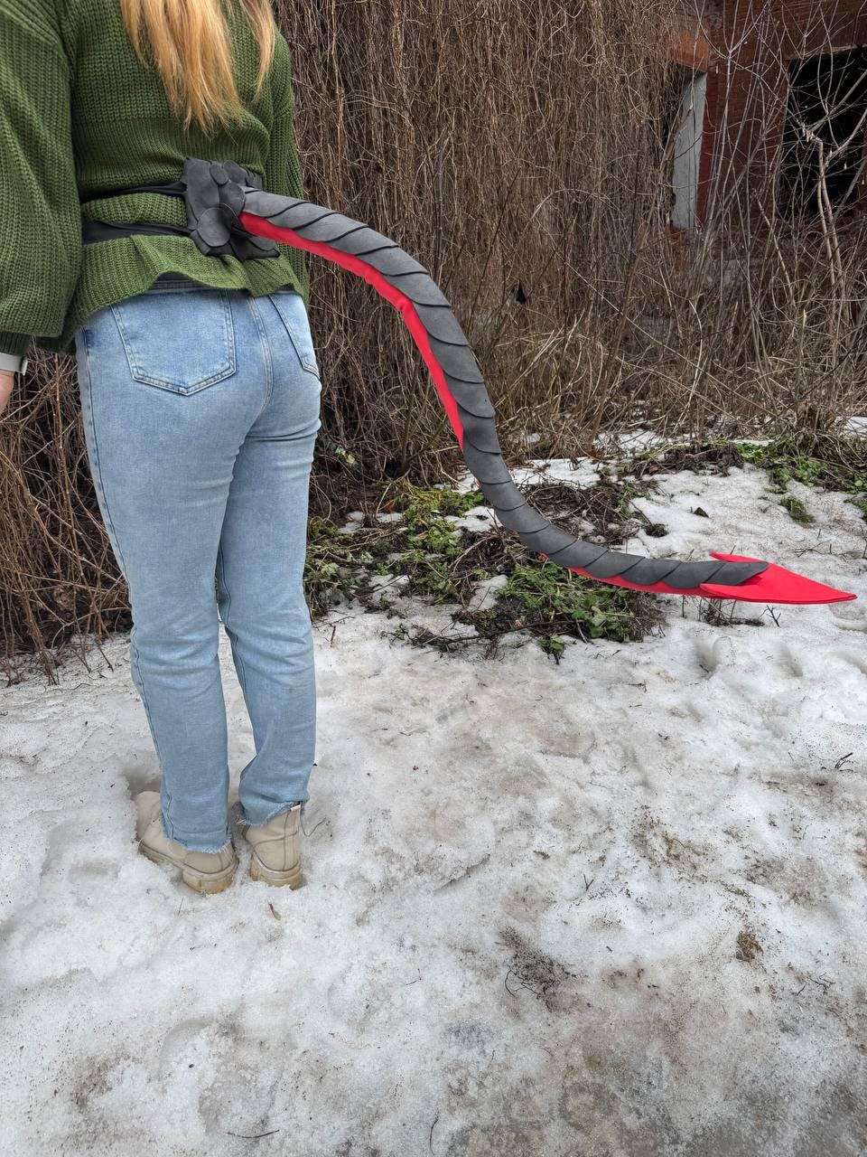 May include: A gray and red costume tail attached to a black belt. The tail is curved and has a scale-like pattern. The tail is attached to a black belt worn around the waist. The tail is red at the tip.