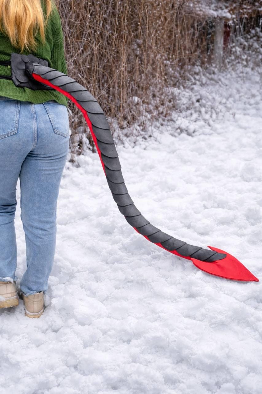 May include: A black and red dragon tail costume accessory. The tail is made of a segmented, textured material with a red underside and a pointed tip. The tail is attached to a black belt.