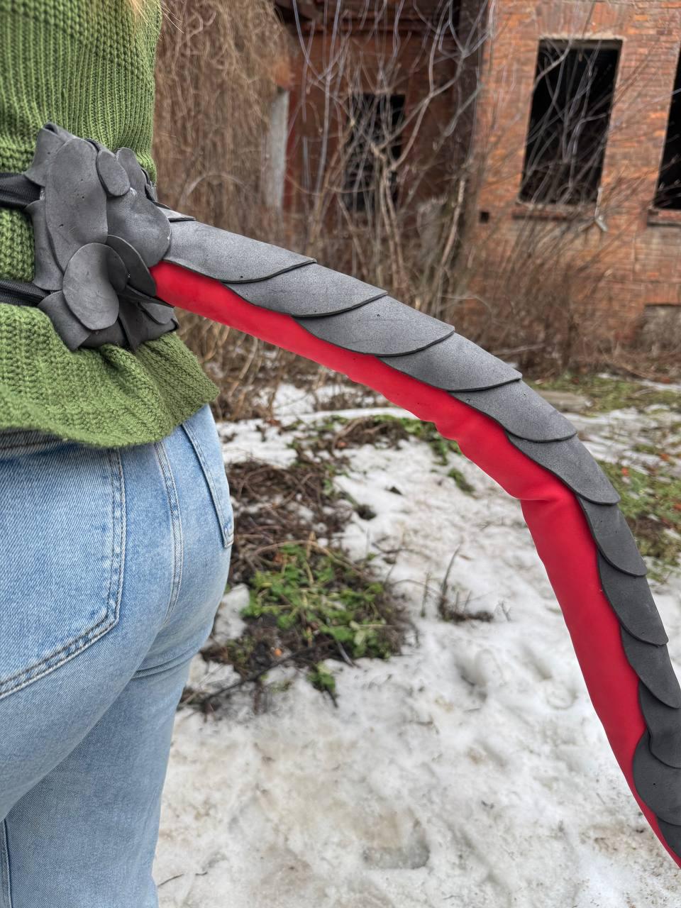 May include: A gray and red dragon tail prop, featuring a layered scale design. The tail is attached to a black belt. The prop is made of foam and fabric. The tail is red with gray scales.