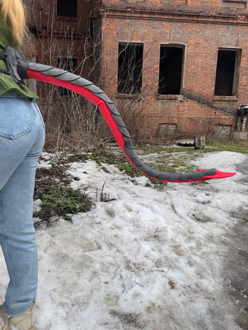 May include: A red and gray dragon tail prop, with a black spine, is held by a person wearing a green sweater and blue jeans. The tail is made of fabric and is curved. The background includes a brick building and snow.