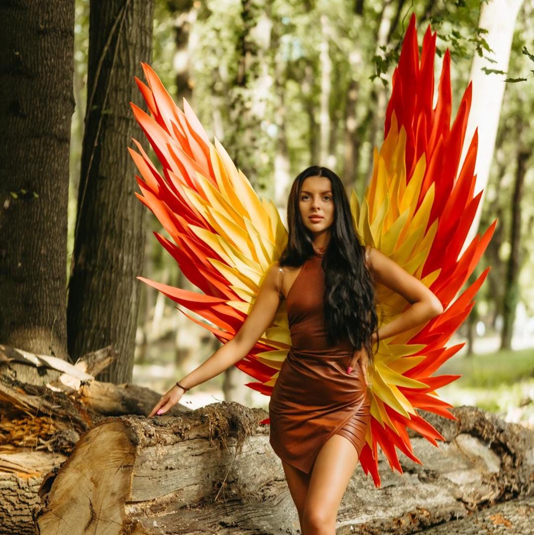 May include: Large decorative wings featuring a vibrant gradient of red, orange, and yellow pointed feathers. The wings are worn as a back accessory, extending upwards and outwards with a structured, layered design that mimics a flame-like appearance.