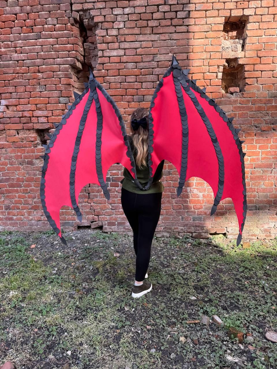 May include: Red dragon wings with black trim and accents. The wings are spread wide, showcasing their vibrant color and design. The wings are attached to a person wearing a green top and black pants, standing in front of a brick wall.