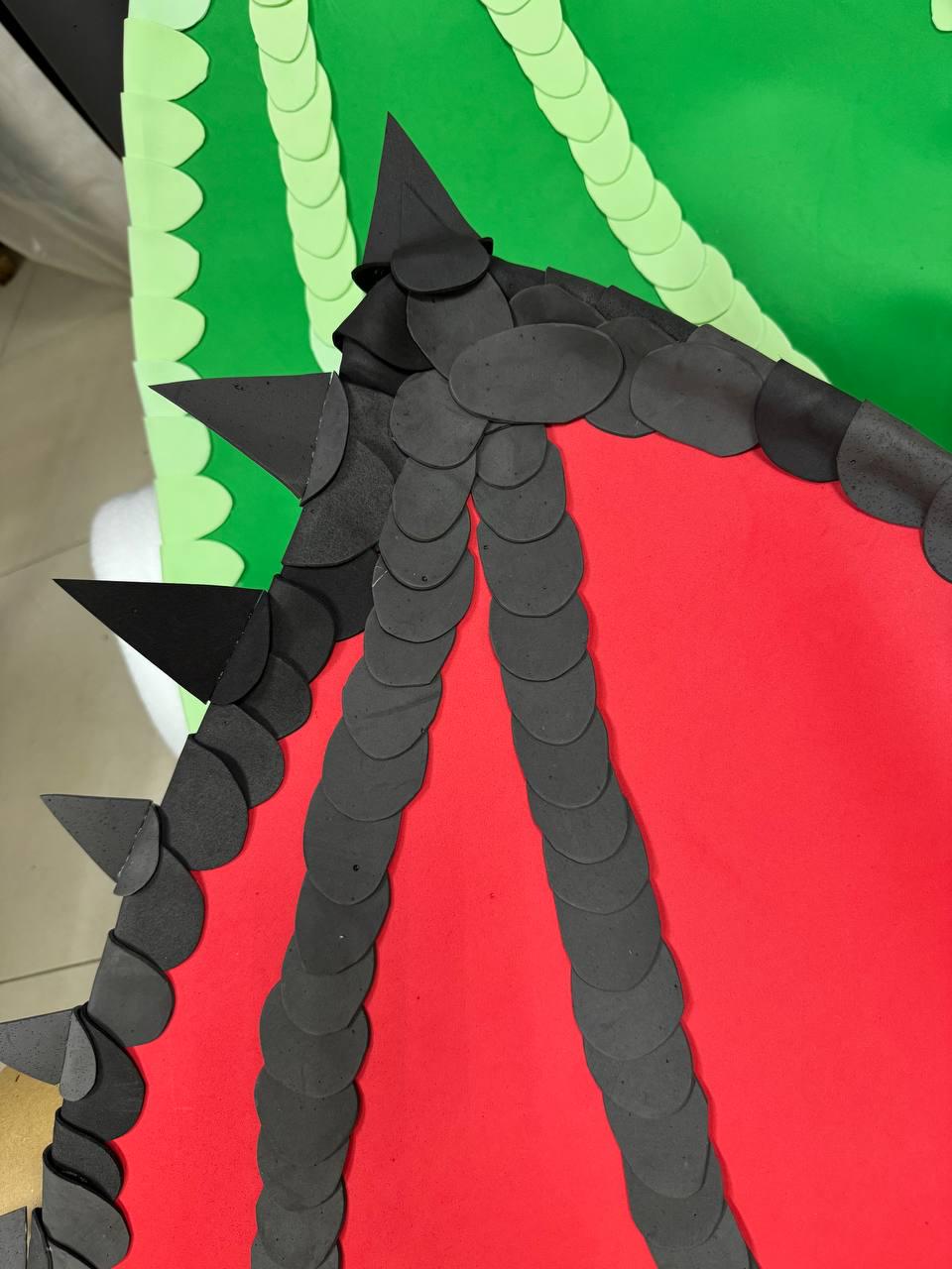 May include: A close-up of a dragon wing made of black, gray, red, and green paper. The wing features black spikes and a pattern of overlapping gray circles. The background is green with a scalloped edge.