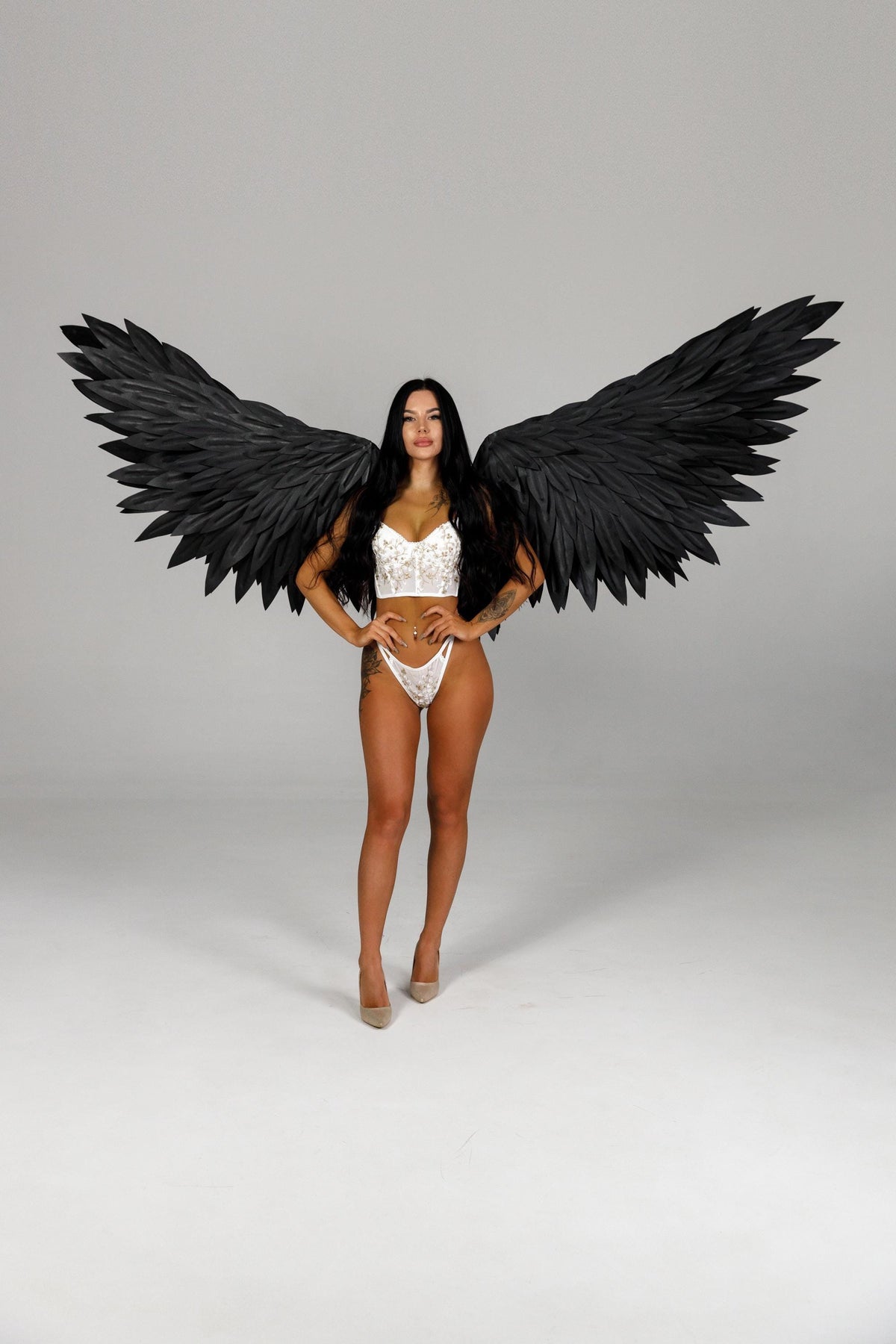 May include: A woman wearing a white lace lingerie set and black feathered angel wings. The wings are large and spread out behind the person. The person is wearing nude high heels.