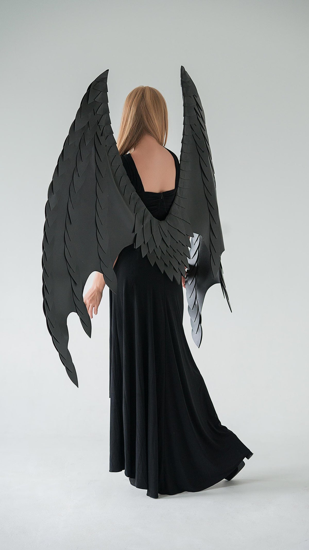 May include: A person wearing a long black dress and large, black, layered costume wings. The wings have a detailed, feathered appearance and are spread out behind the person. The background is a plain, light gray.