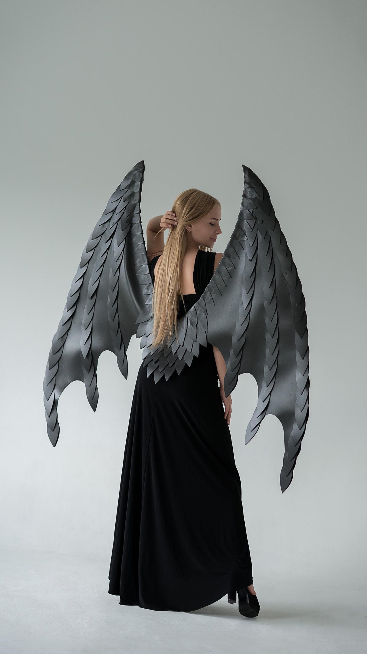 May include: Large, gray dragon wings with a detailed, layered design. The wings are worn by a person in a black dress. The wings have a textured appearance and are spread wide, creating a dramatic silhouette against a white background.