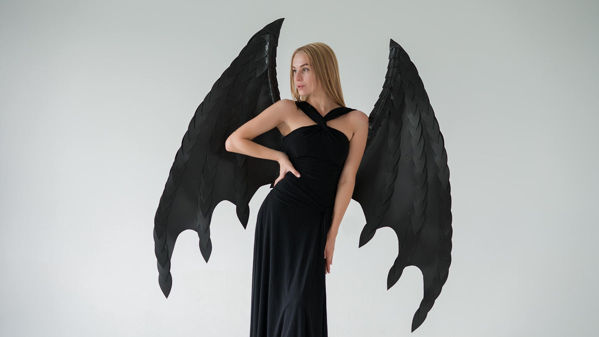 May include: A woman wearing a black dress and large black costume wings. The wings have a textured, layered design and are spread out behind the person. The dress is a simple, elegant style.