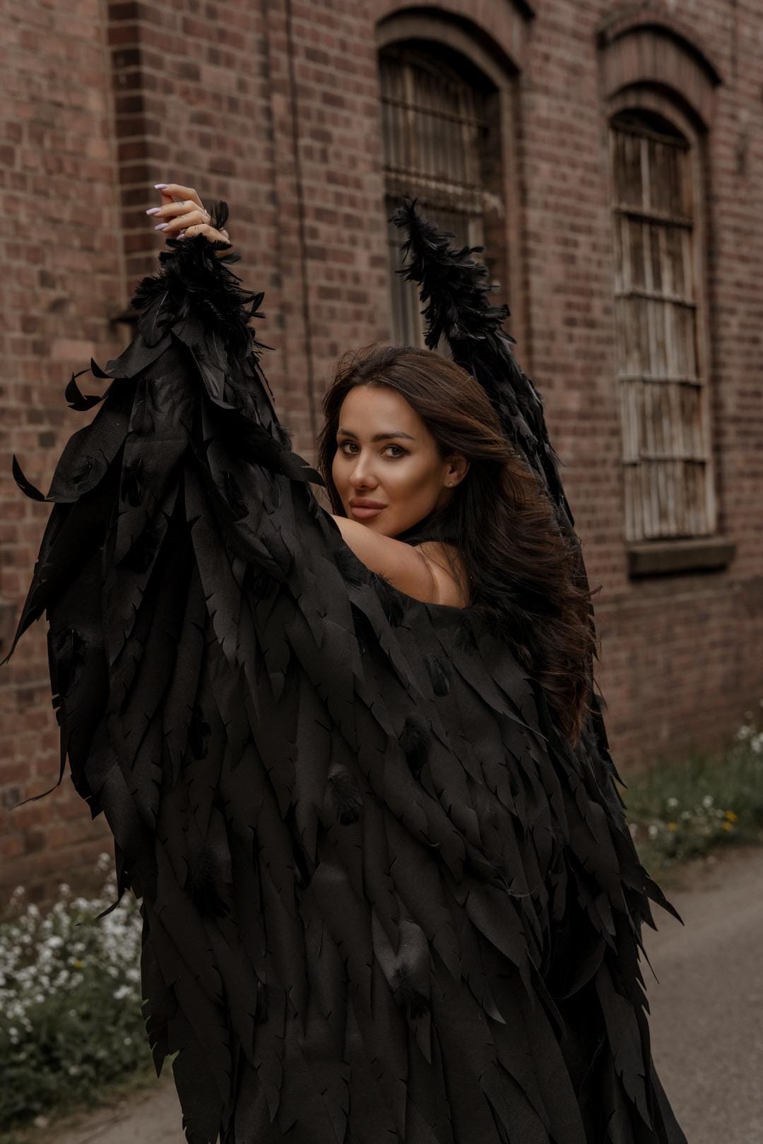 May include: A person wearing large, black feather wings, posing outdoors. The wings are the focal point, with each feather detailed. The background features a brick building and a pathway, creating a dramatic contrast with the dark wings.