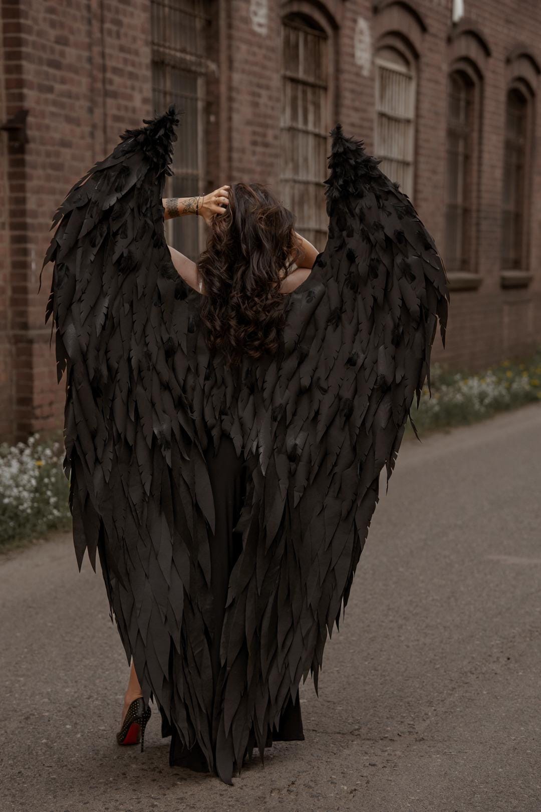 May include: A person wearing large, black feathered angel wings, standing on a paved road. The wings are spread wide, creating a dramatic silhouette against a brick building backdrop. The person is wearing a black dress and high heels.