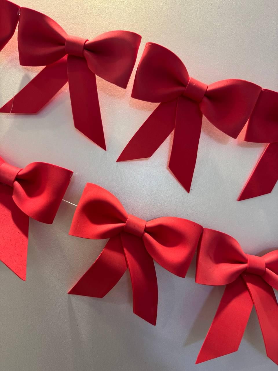 May include: A string of bright red decorative bows, each with a classic bow shape and long, angled tails. The bows are evenly spaced and appear to be made of a matte material. The background is a neutral, off-white color.