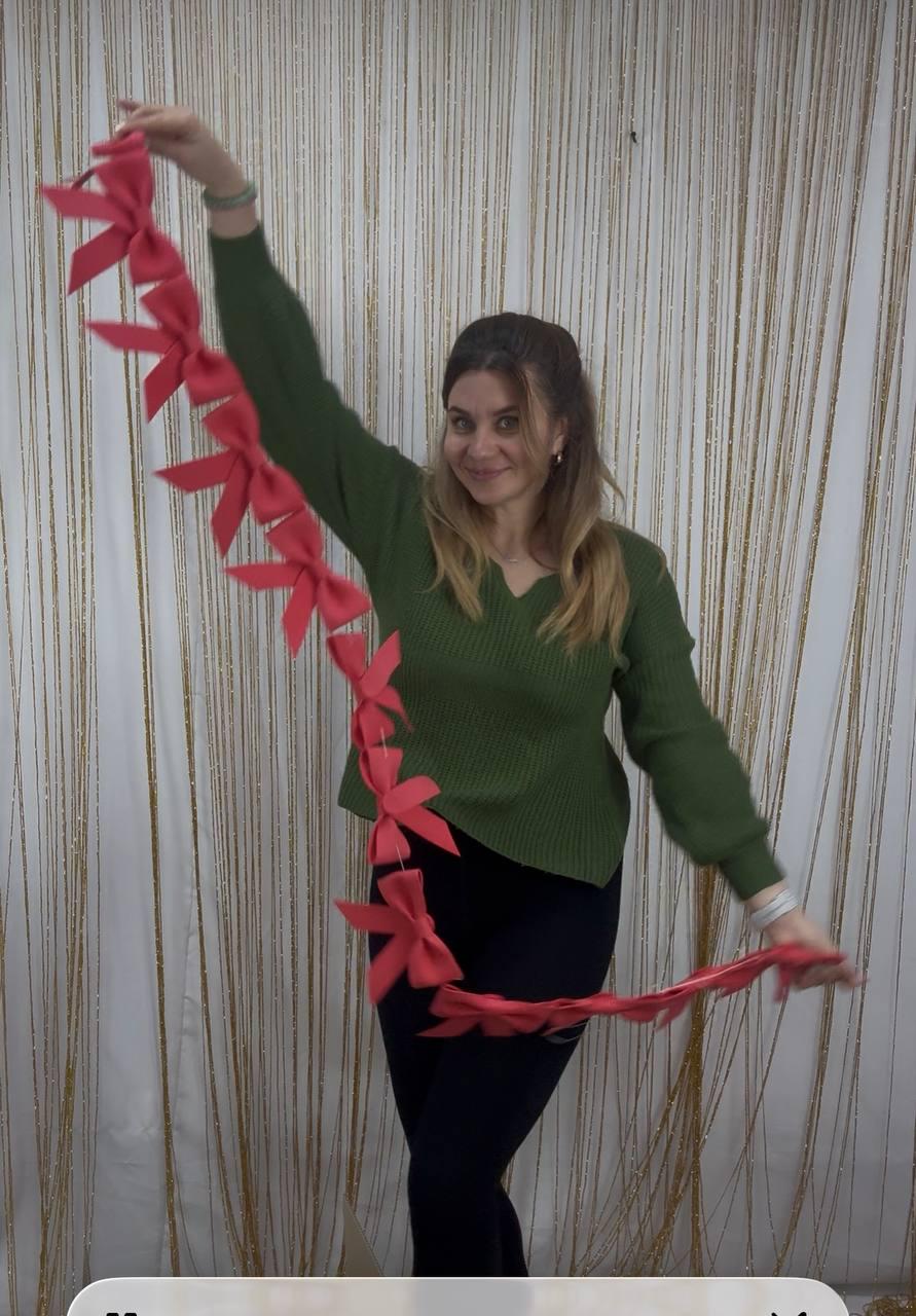 May include: A woman holds a garland of red bows. The bows are evenly spaced along a red ribbon. She is wearing a green sweater and black pants. The background is a gold-colored curtain.