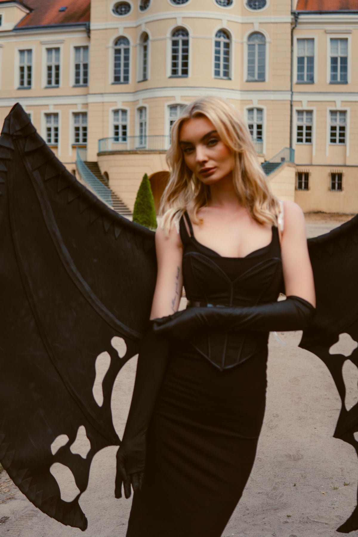 May include: A black dress with a corset-style bodice and long black gloves. Large black wings with a decorative pattern are behind the dress. The background features a light-colored building with multiple windows.
