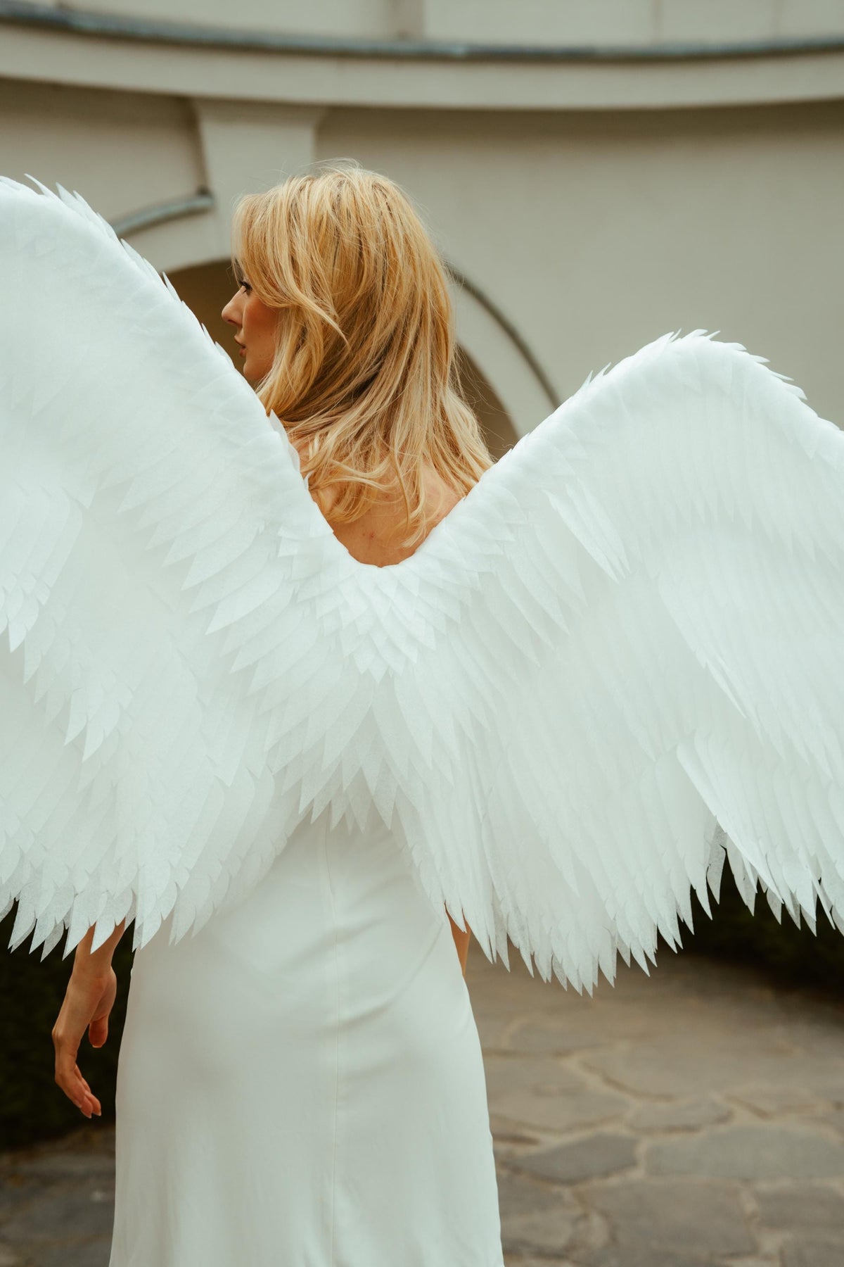 May include: A person wearing a white dress and large, feathered angel wings. The wings are spread wide, with each feather individually detailed. The person has blonde hair and is seen from the back, with a blurred background.