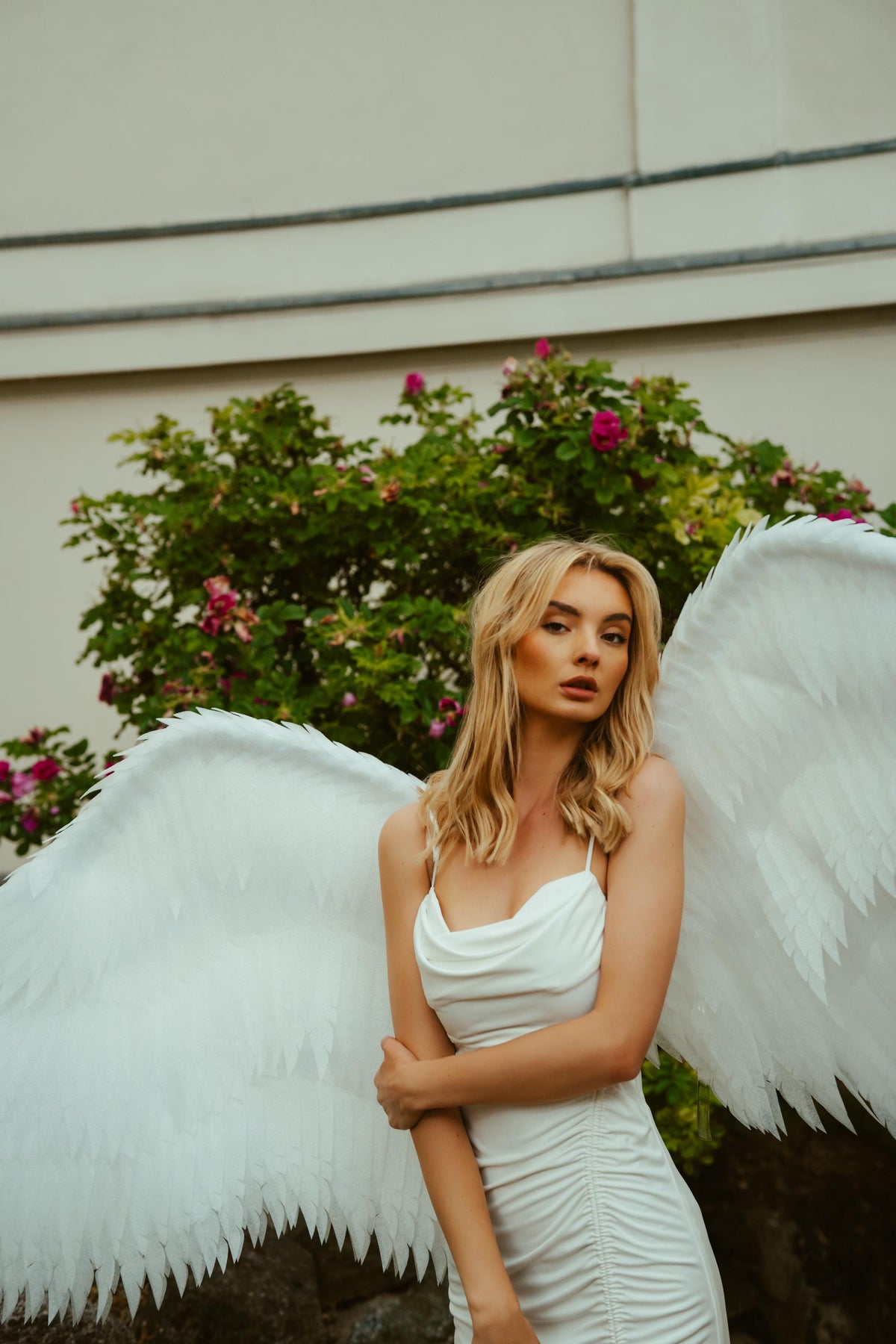 May include: A woman wearing a white dress and large white angel wings stands in front of a green bush with pink flowers. The dress has spaghetti straps and a ruched design.