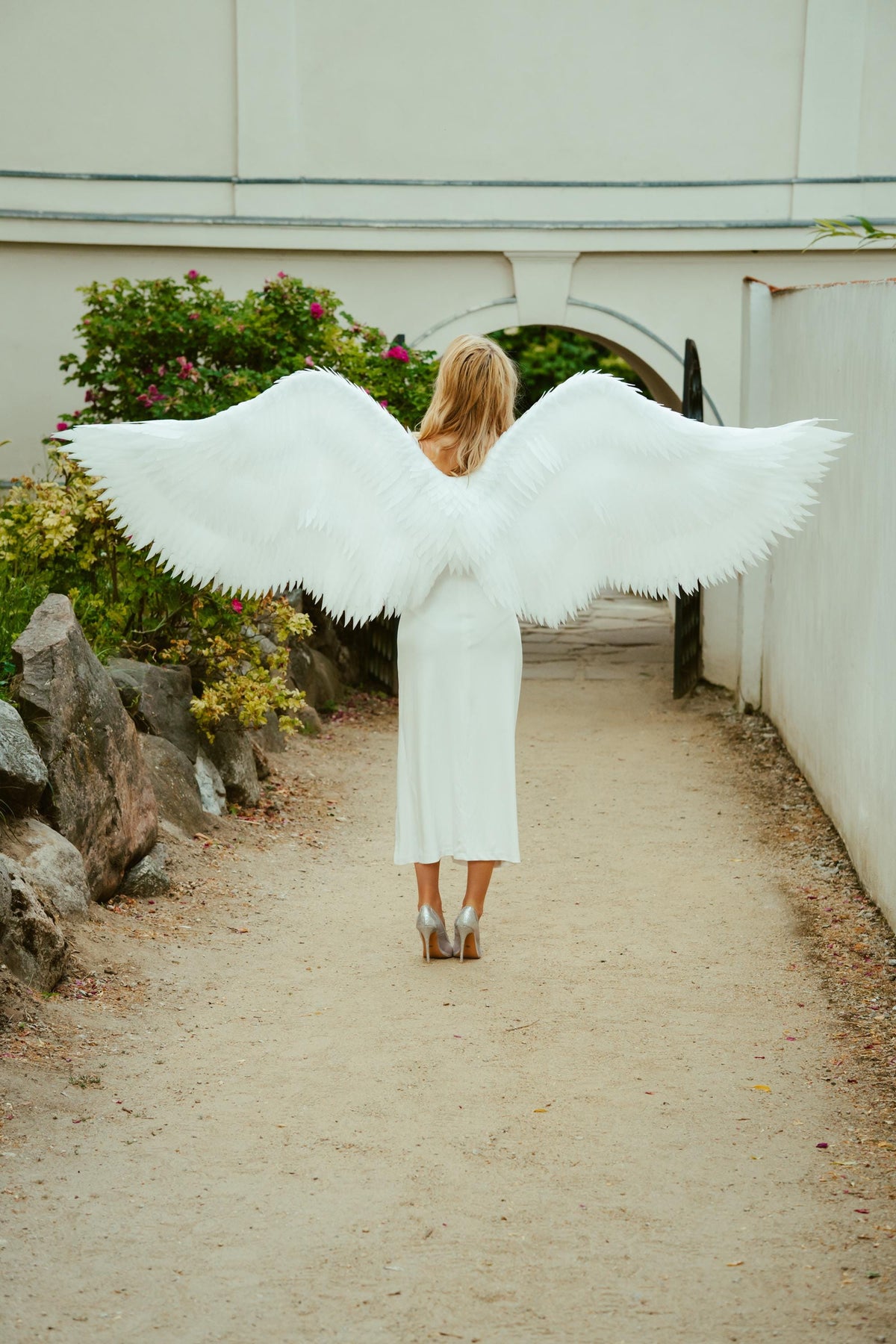 May include: A person wearing a white dress and silver heels, with large white feathered angel wings. The person is standing on a dirt path, with a stone wall and greenery on the left and a white wall on the right.