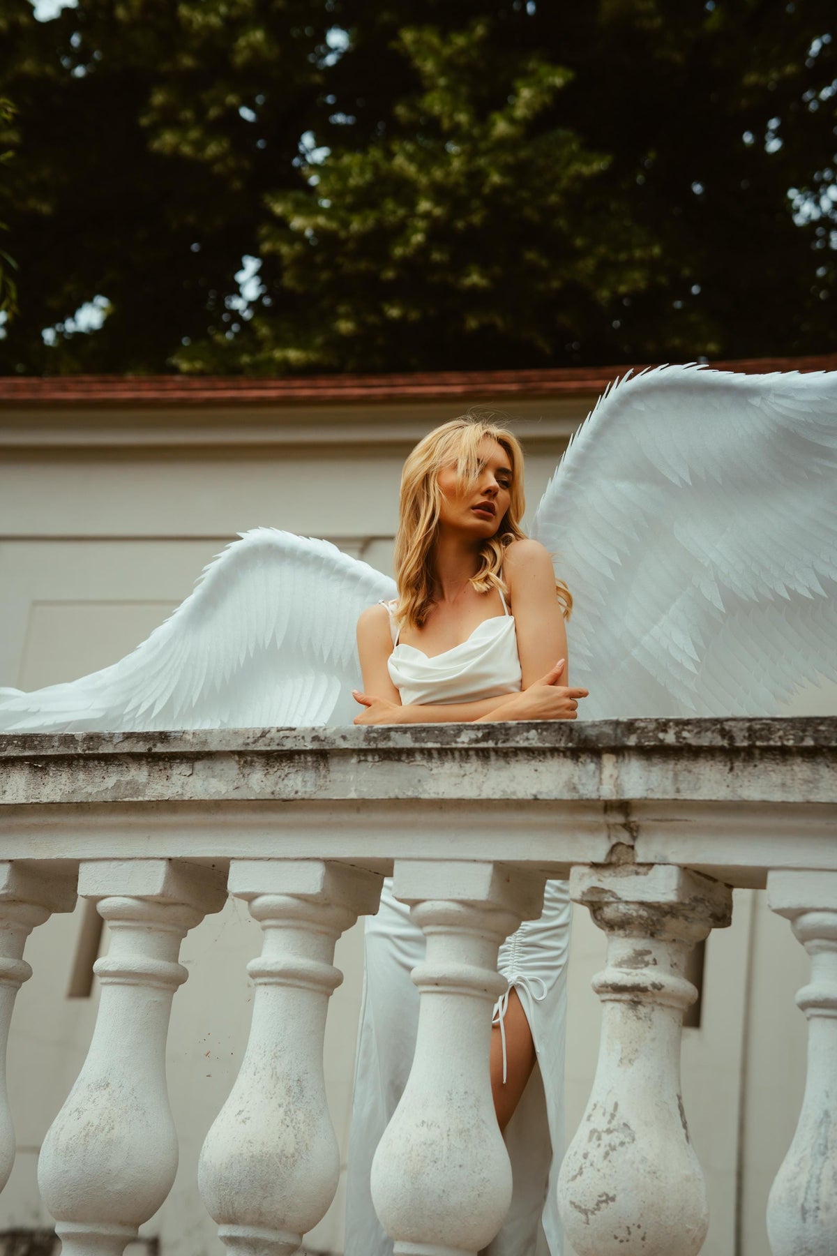 May include: A woman wearing a white dress and large white angel wings, leaning on a white stone balustrade. The background features a green tree line and a white building. The dress has a cowl neckline and a side slit.