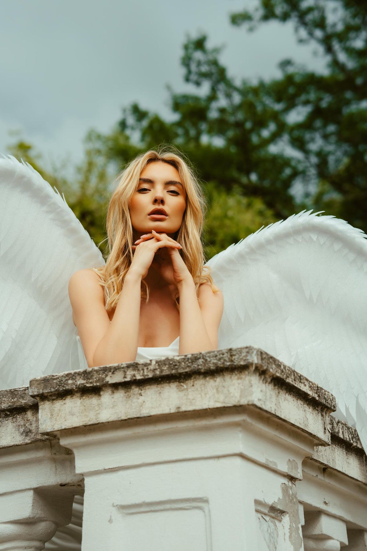 May include: A person with long blonde hair and white angel wings poses outdoors. The person is wearing a white dress and resting their hands on a stone ledge. The background features green trees and a cloudy sky.
