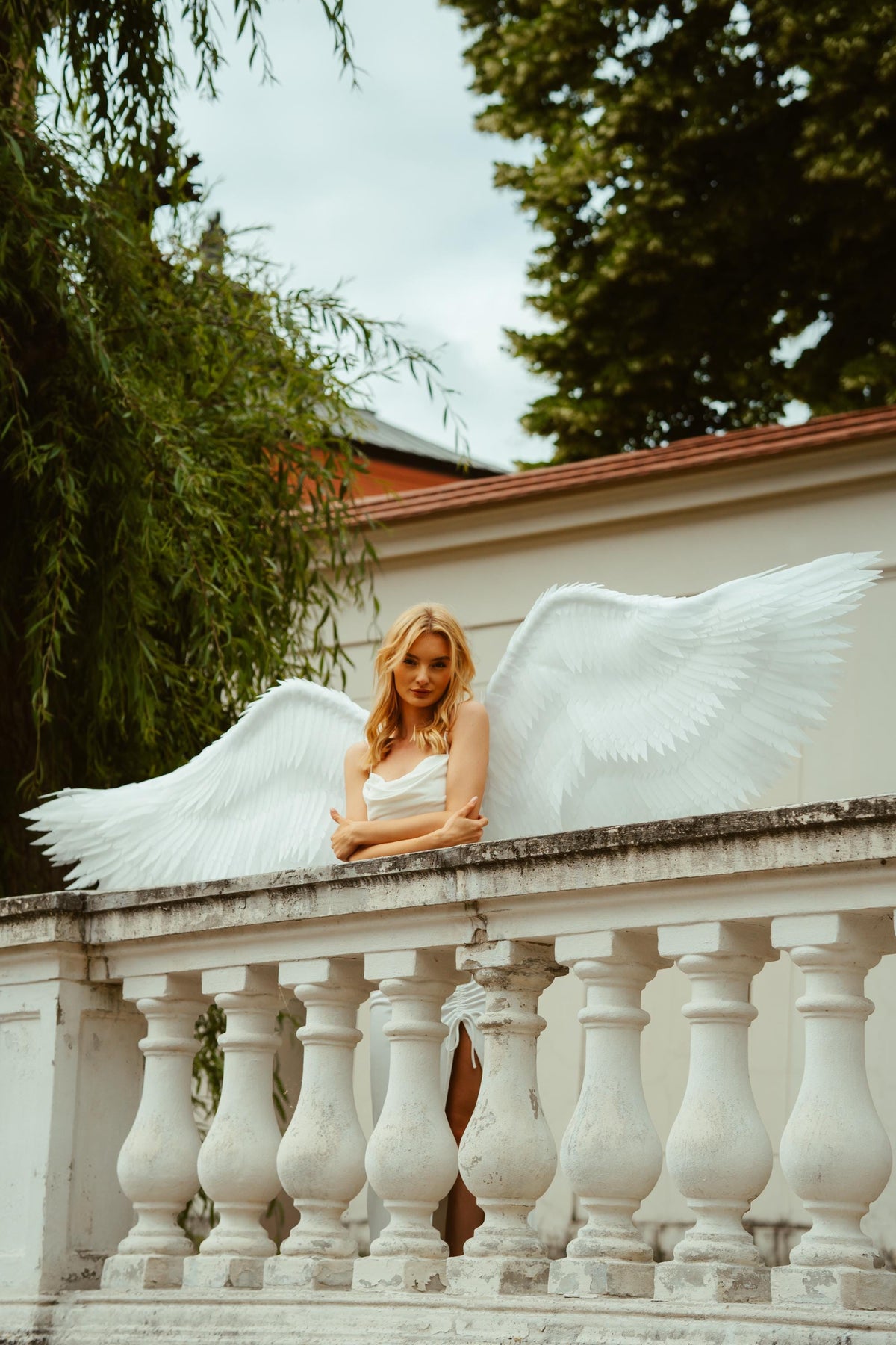 May include: A woman with long blonde hair wearing a white dress and large white angel wings, leaning on a white stone balustrade. The wings are spread wide, and the background includes trees and a building.