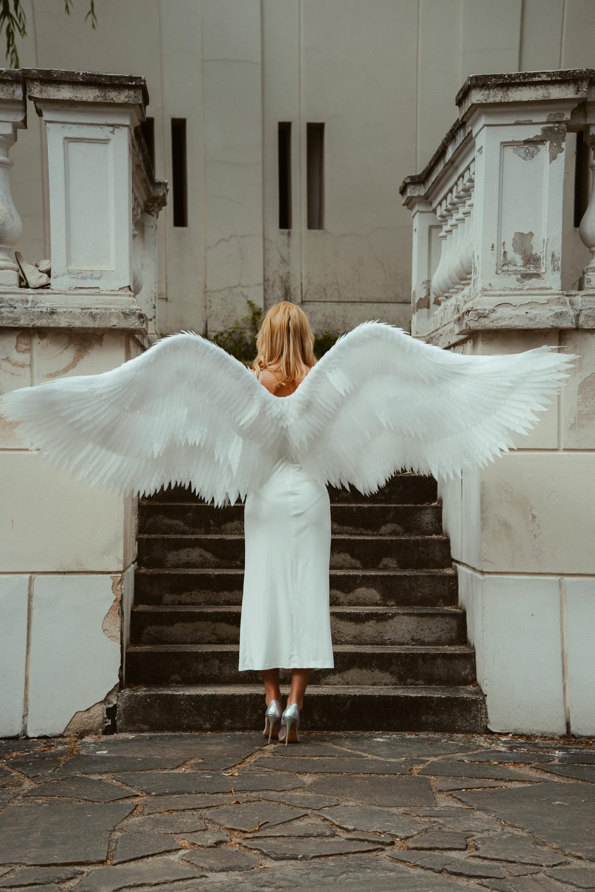 May include: A person wearing a white dress and silver heels, with large white feathered angel wings. The person is standing on a stone staircase in front of a white building with architectural details.