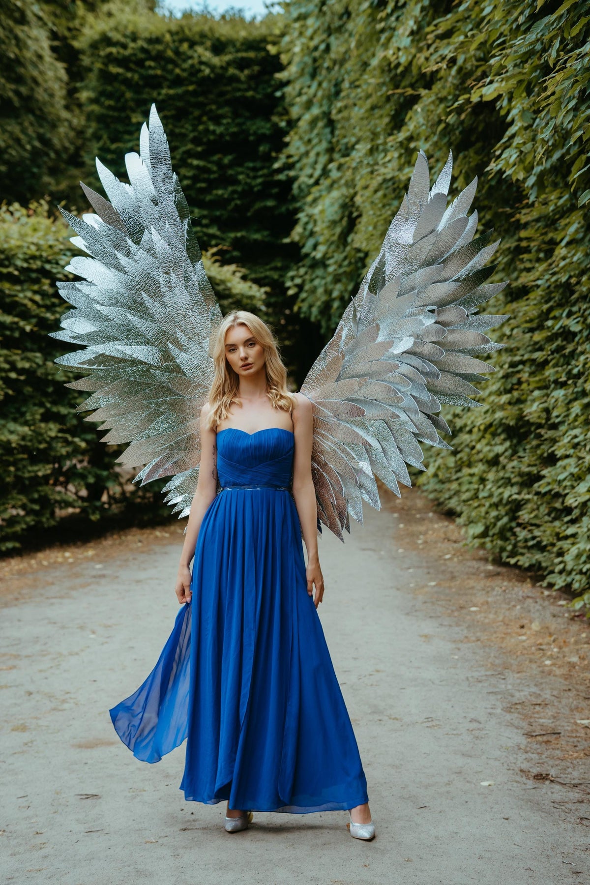 May include: A woman wearing a strapless, royal blue gown and large silver angel wings stands on a path. The wings are made of multiple layers of reflective material. The dress has a flowing skirt and a fitted bodice. The background is a lush green park.